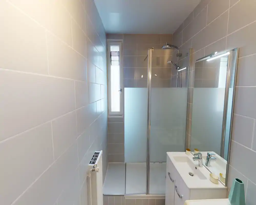 Baño moderno y compacto con ducha a ras de suelo con vidrio esmerilado, mueble lavabo con espejo y grifería cromada, baldosas grises neutras que aportan un aspecto limpio y contemporáneo.