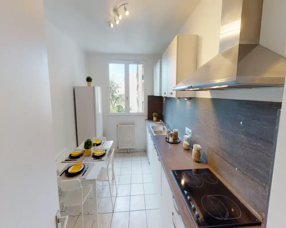 Cocina moderna y luminosa en galería con una encimera larga, placa de inducción, campana de acero inoxidable y una pequeña mesa para cuatro junto a la ventana.