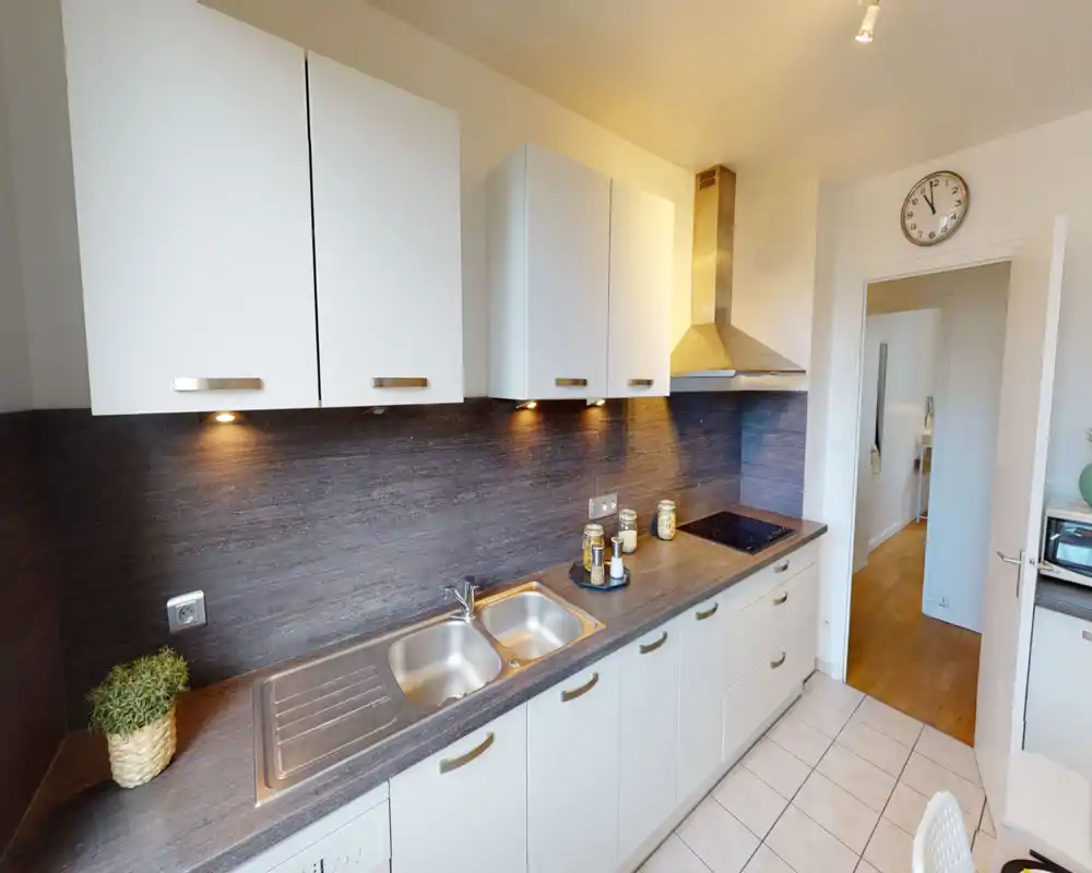 Cocina moderna y luminosa con armarios blancos, fregadero de acero inoxidable y placa de cocina integrada; luces bajo los muebles y tarros decorativos crean un espacio ordenado y funcional.