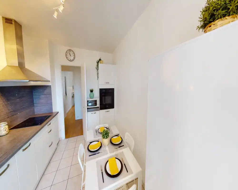 Cocina moderna y luminosa con horno integrado, placa de cocción y una pequeña mesa para cuatro. Muebles blancos, encimera efecto madera y plantas decorativas aportan un ambiente limpio y acogedor.