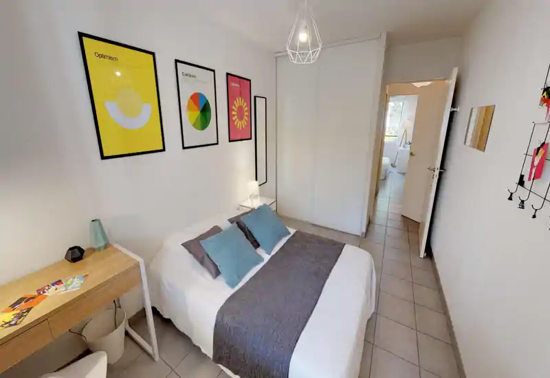 Bright, stylish bedroom with a double bed dressed in white linen, decorative pillows and a grey runner. The room features framed colorful prints, a small desk, built-in wardrobe with sliding doors and clean tiled flooring — ideal for a cozy stay.