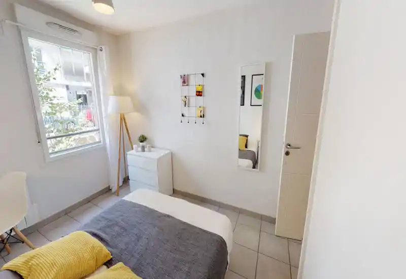 Bright, modern bedroom with a neatly made bed, soft textiles and a floor lamp by the window. Large window provides natural light and a simple dresser with decorative accents completes the space.