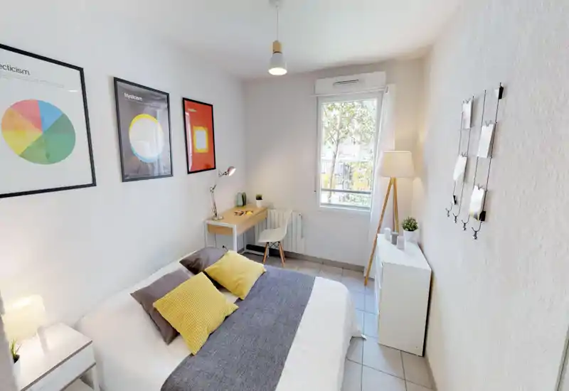 Bright, modern bedroom with a double bed dressed in white linens, yellow accent cushions and a gray runner; a small desk by the window and tasteful framed artwork along the wall create a cozy, well-lit space.