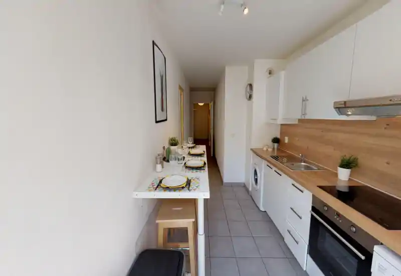Narrow modern kitchen with wooden countertops, white cabinets and a breakfast bar set for several guests; compact layout includes oven, sink and washing machine.