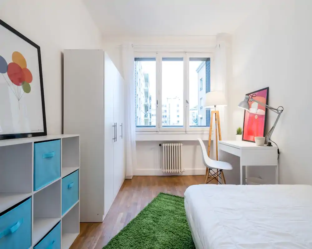 Bright, modern bedroom with a comfortable bed, white furnishings and a study corner by the window. Large window floods the room with natural light; minimalist décor and a green rug create a fresh, welcoming atmosphere.