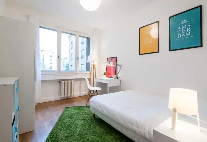 Bright, minimalist bedroom with a comfortable double bed, green rug, desk and large window letting in abundant natural light. Simple, modern décor with framed prints and bedside lamp create a calm, homey atmosphere ideal for rentals.