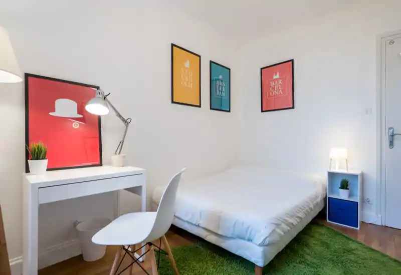 Bright, modern bedroom with a neatly made double bed, a small desk and chair, colorful framed prints and soft lighting — cozy and well-styled for a short stay.