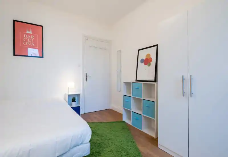 Bright, minimal bedroom with a neatly made bed, green rug and simple storage units; white walls and colorful accents give a fresh, airy feel.