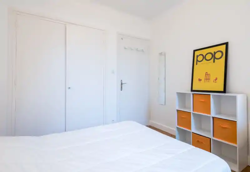 Bright, minimal bedroom with a neatly made white bed, built-in wardrobe doors and a shelving unit with orange storage bins adding a playful accent.