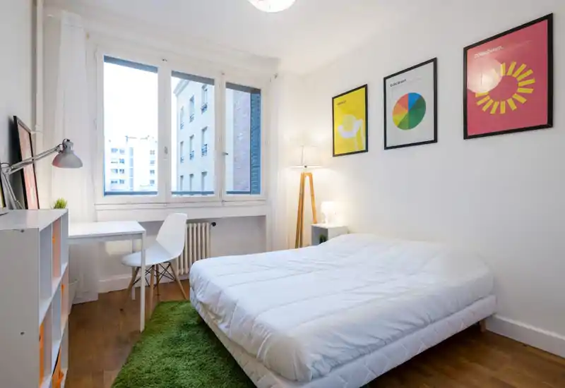 Bright, modern bedroom with a large window, simple white bedding and warm decorative touches including framed prints and a green rug.