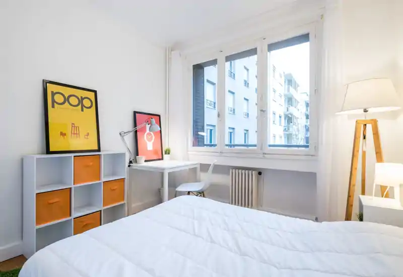 Bright, modern bedroom with a large window, white bedding and a minimal desk area — clean lines, warm lamp lighting and orange accents create a fresh, inviting space.