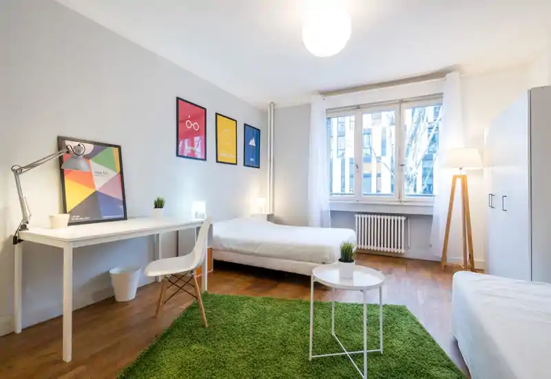 Bright, modern bedroom with a double bed, desk and cozy green rug. Large window provides ample natural light; simple, Scandinavian-style furnishings and soft textiles create an inviting atmosphere for guests.