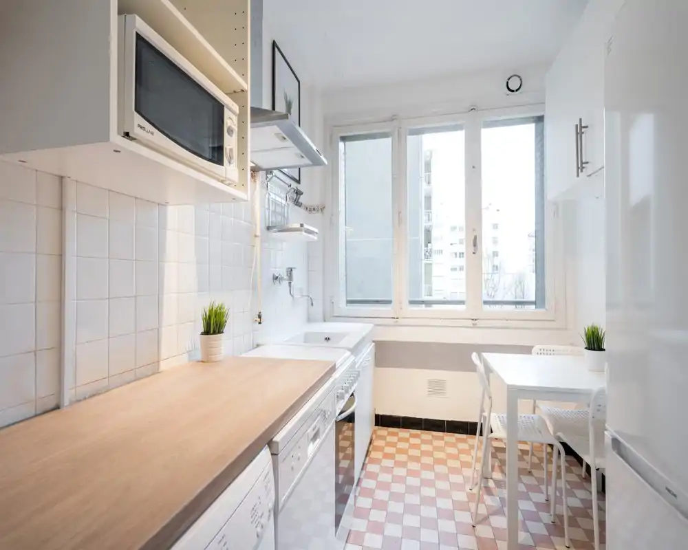 Bright, compact kitchen with a wooden countertop, built-in appliances, tiled backsplash and a small dining table by a large window — clean, functional and airy.