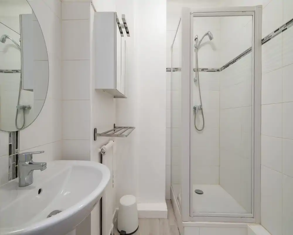Clean, compact bathroom with a corner shower cubicle, wall-mounted mirror cabinet, sink and chrome fixtures; bright white tiles and a small towel rack.