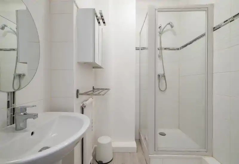 Salle de bains compacte et propre avec cabine de douche d'angle, armoire-miroir murale, lavabo et robinetterie chromée ; carrelage blanc lumineux et petit porte-serviettes.