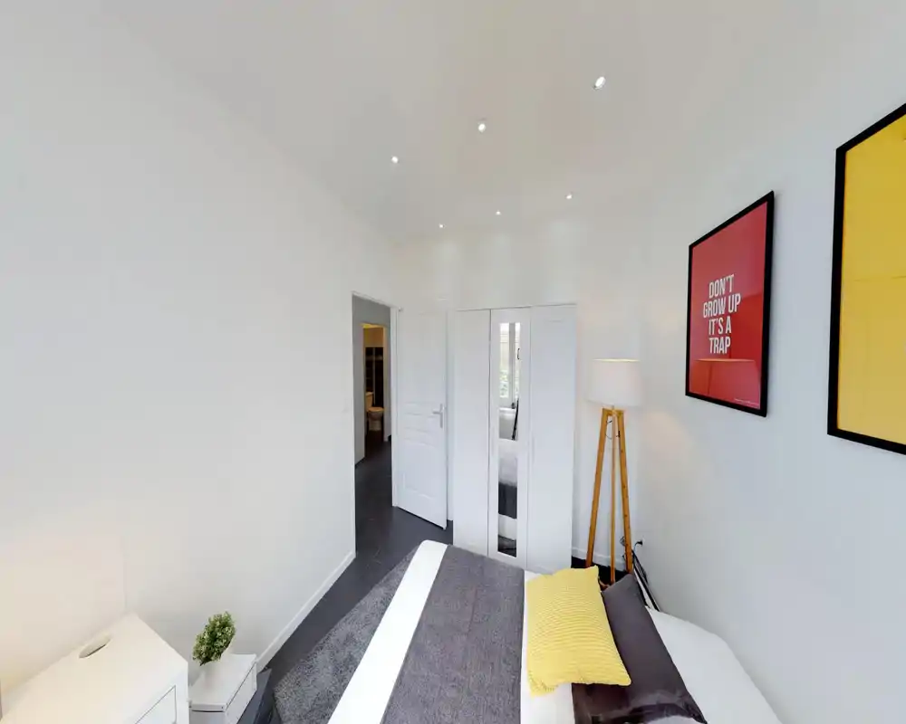 Camera da letto moderna e luminosa con un letto matrimoniale ordinato, coperte accoglienti e una lampada da terra. Pareti bianche, quadri incorniciati e un armadio con specchio donano un aspetto pulito e invitante.