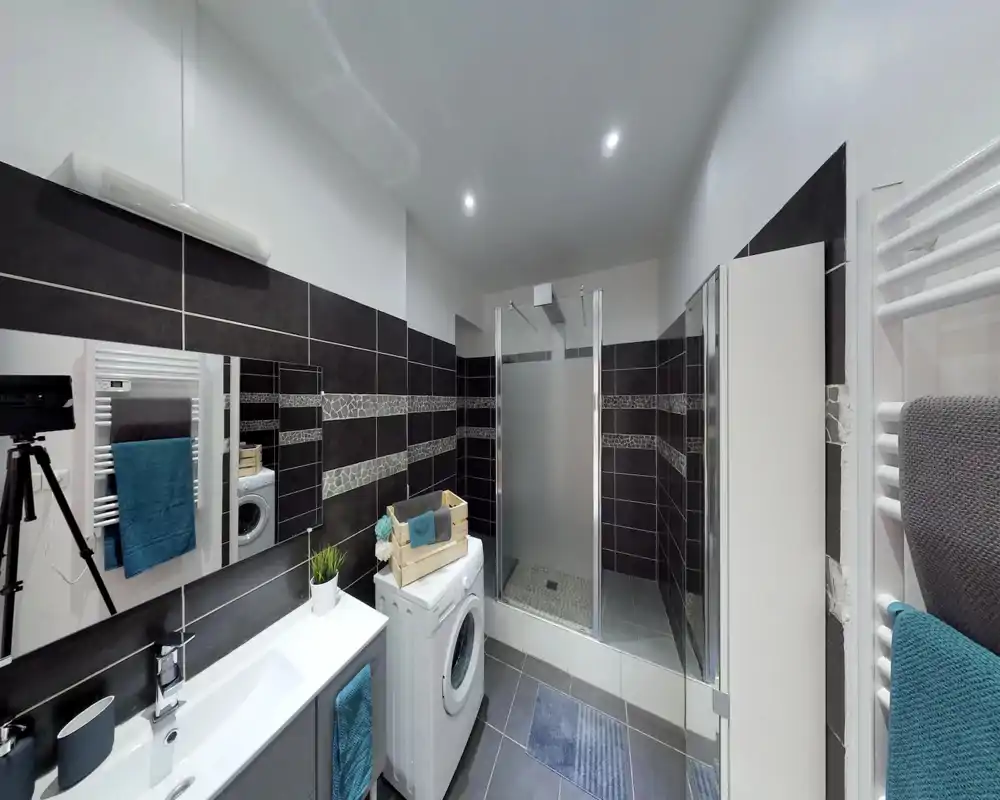 Salle de bains moderne carrelée avec cabine de douche en verre, lavabo, lave-linge et sèche-serviettes ; espace propre et bien éclairé, adapté aux annonces locatives.