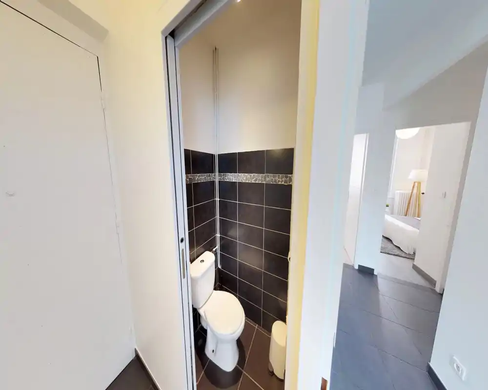 Petite salle d'eau avec WC moderne et carrelage mural sombre ; toilette fonctionnelle et propre attenante au couloir.