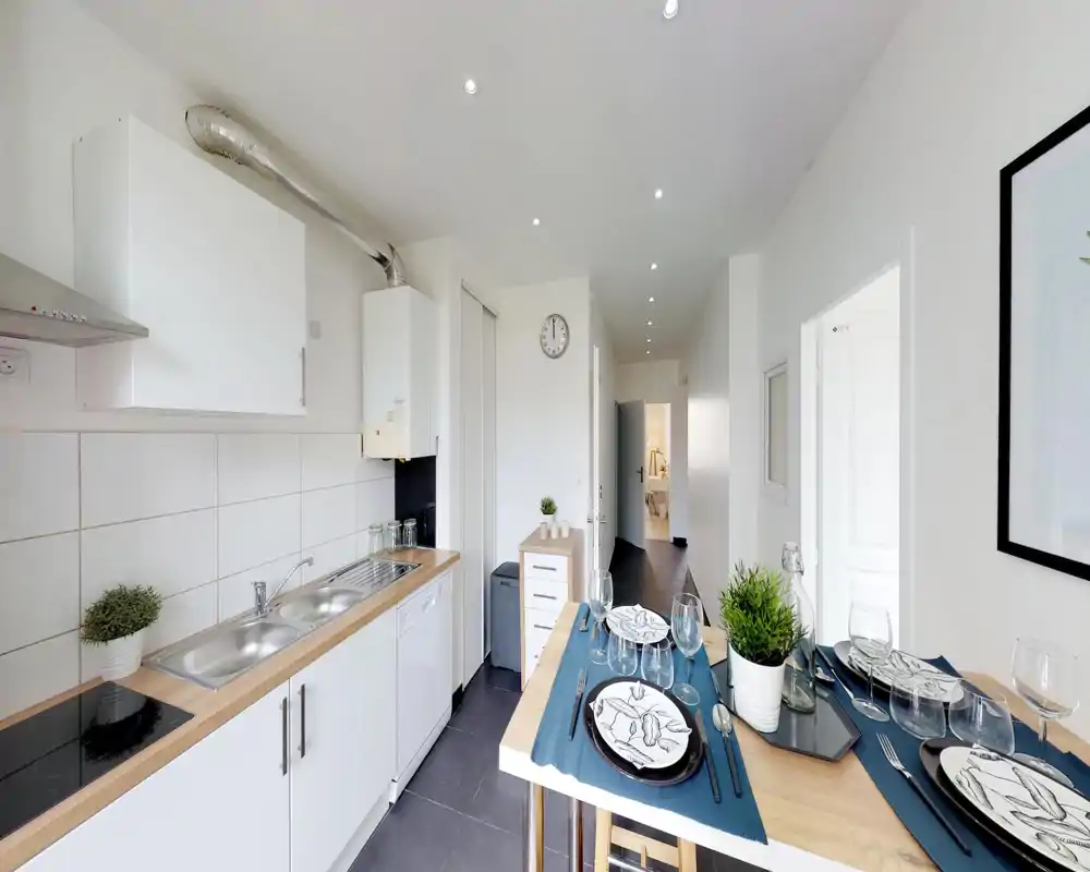 Cuisine moderne et lumineuse avec évier intégré, plaque à induction et petit bar repas pour quatre ; plans de travail en bois clair et meubles blancs pour un espace propre et frais.
