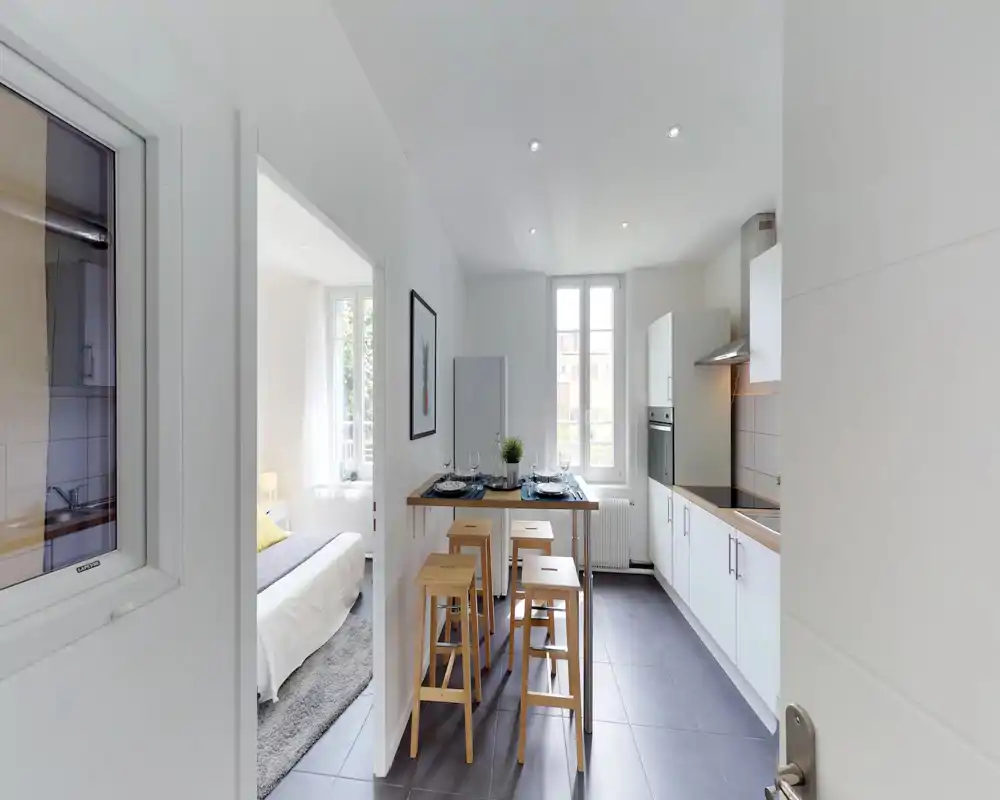 Cuisine moderne et lumineuse avec meubles blancs, four encastré et une table haute avec tabourets en bois — idéale pour repas décontractés.