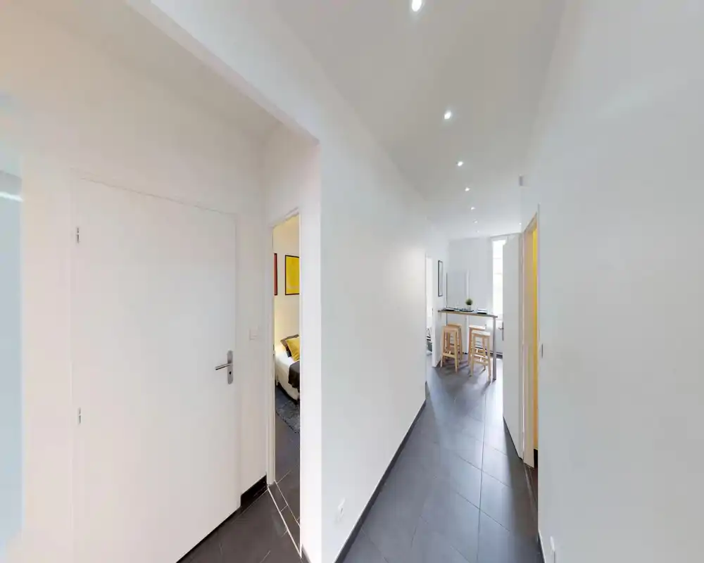 Couloir lumineux et minimaliste avec sol carrelé menant à la cuisine et aux pièces latérales ; finitions modernes et spots encastrés pour une entrée nette.