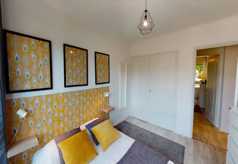 Bright, styled bedroom with a patterned yellow headboard, two mustard cushions and a modern pendant lamp; the bed is clearly visible and the room feels warm and well-lit.