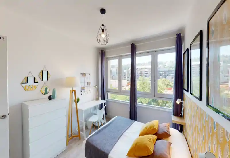 Sunny, modern bedroom with a comfortable double bed, mustard-yellow accent pillows, a gray runner and large windows offering a bright city view. The room features a small desk, dresser and tasteful wall art, creating a cozy, well-lit space ideal for short-term rentals.