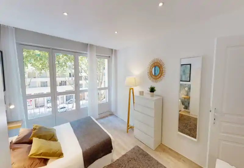 Bright, modern bedroom with a large window, neatly made double bed, warm textiles and a dresser with a standing lamp — ideal for comfortable stays.