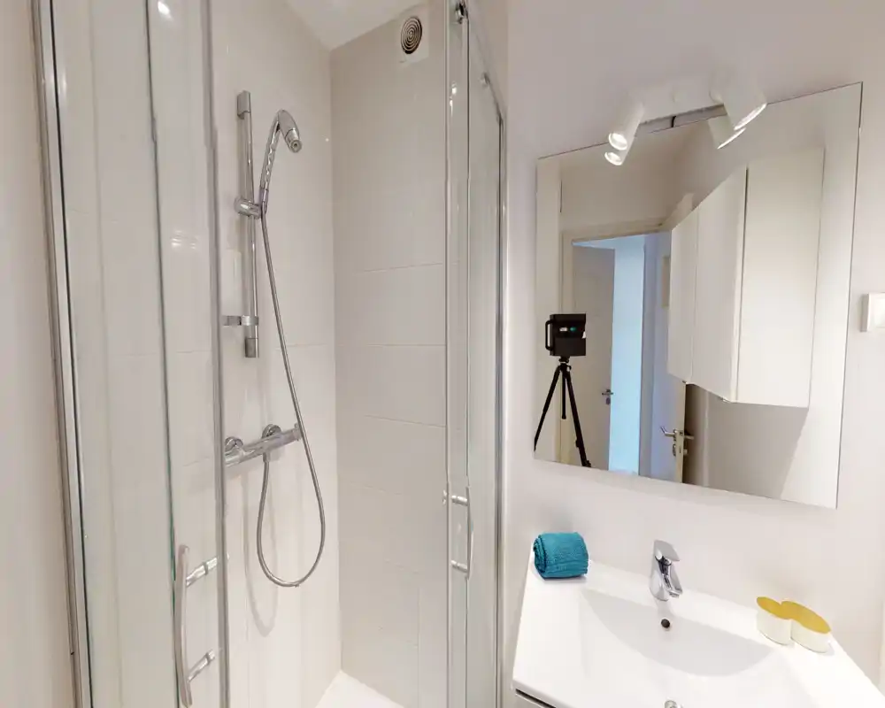 Clean, modern bathroom with a glass shower enclosure, handheld shower head, white tiles and a sink with mirror — bright and well-maintained.
