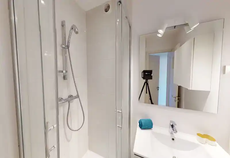 Salle de bain moderne et propre avec cabine de douche en verre, pommeau de douche manuel, carrelage blanc et lavabo avec miroir — lumineux et soigné.