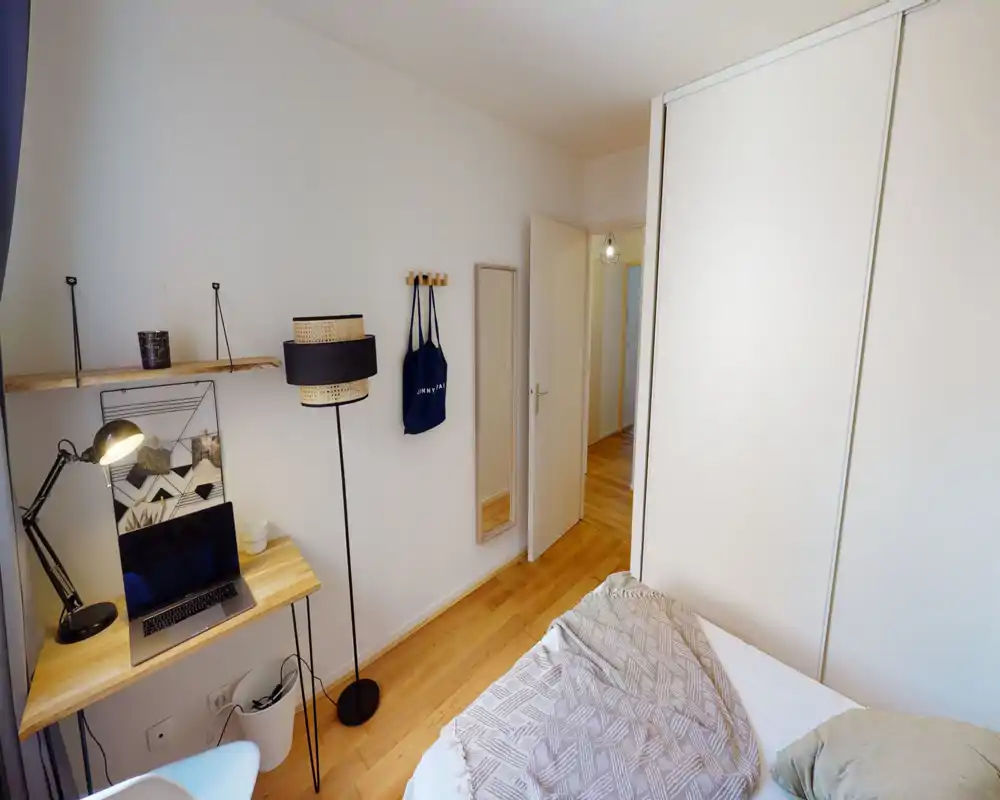 Compact, bright bedroom with a double bed, warm wooden floor and a small workspace with a laptop. Clean minimalist decor, wardrobe with sliding doors and a full-height mirror near the entrance.