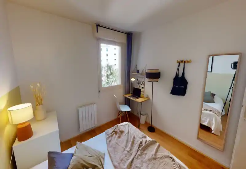 Bright, tidy bedroom with a comfortable bed in the foreground, warm bedside lamp and a small workspace by the window — ideal for relaxing or remote work.
