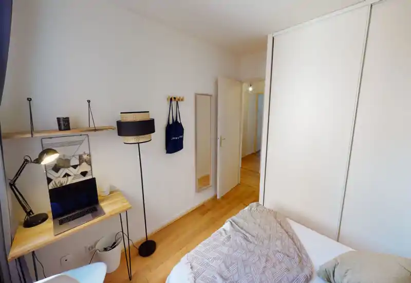 Compact, bright bedroom with a double bed, warm wooden floor and a small workspace with a laptop. Clean minimalist decor, wardrobe with sliding doors and a full-height mirror near the entrance.
