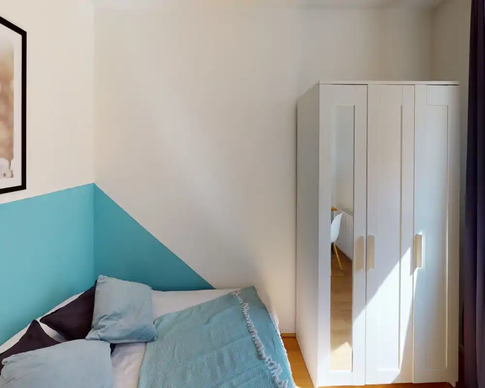 Camera da letto luminosa e ordinata con parete accentata turchese, letto visibile con cuscini e coperta coordinata e un armadio bianco con pannello a specchio che riflette parte della stanza.