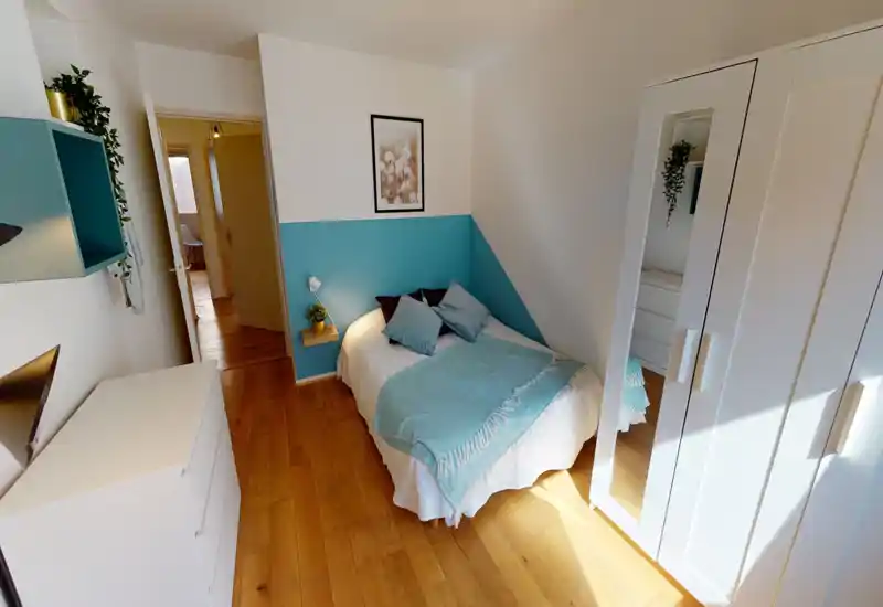 Bright, neatly staged bedroom with a double bed, soft blue accents and natural wood flooring. The space feels welcoming and well lit, with a large wardrobe and dresser for storage.