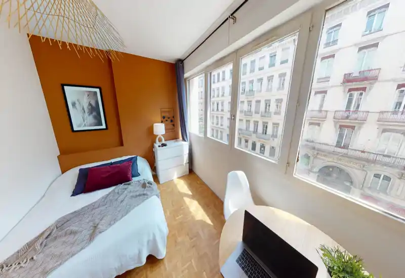 Bright, stylish bedroom with a comfortable double bed set against a warm ochre accent wall, large windows letting in abundant daylight, a small dresser and a workspace by the window.