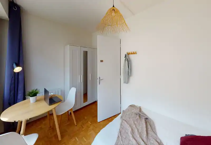 Bright, minimalist bedroom with a visible double bed draped with a textured throw, a small wooden table with chairs and a wardrobe near the door — airy and simply furnished.