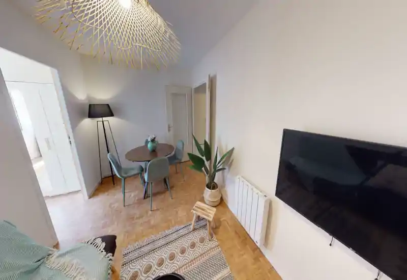 Bright, well-styled living/dining area with a round dining table, four upholstered chairs, decorative rug and wall-mounted TV — cozy and ready for guests.