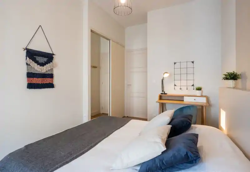 Bright, stylish bedroom with a neatly made bed, decorative cushions and a small wooden desk with lamp — calm neutral tones and soft textiles create a welcoming atmosphere.