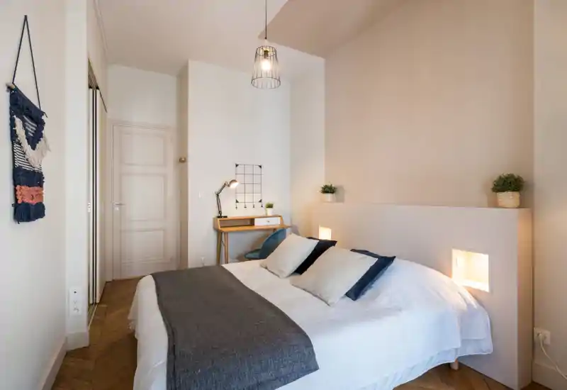 Bright, neatly styled bedroom with a large bed, layered pillows and a textured throw. A small desk and warm recessed lights add practical and cozy touches.