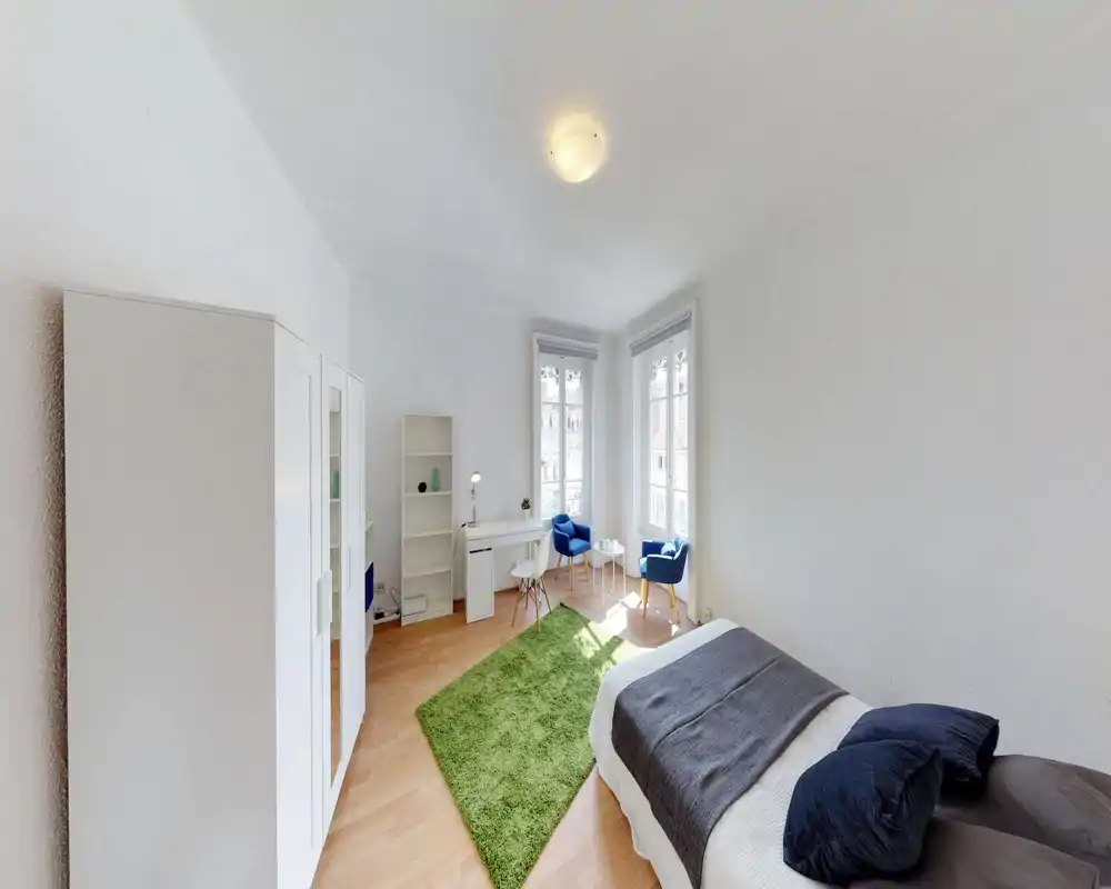 Bright, well-staged bedroom with a comfortable single/double bed in the foreground, a green rug, wardrobe and a cozy sitting area by two tall windows that flood the room with natural light.