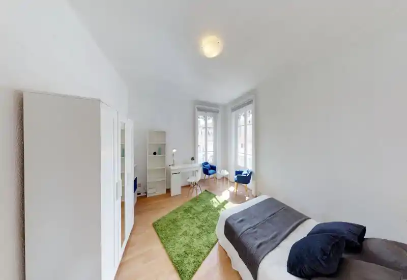 Bright, well-staged bedroom with a comfortable single/double bed in the foreground, a green rug, wardrobe and a cozy sitting area by two tall windows that flood the room with natural light.
