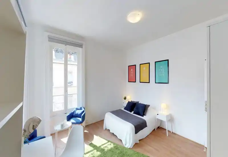 Bright, well-staged bedroom with a double bed, bedside tables and lamps, a comfortable armchair by a large window and colorful framed prints on the wall — ideal for rental photos.