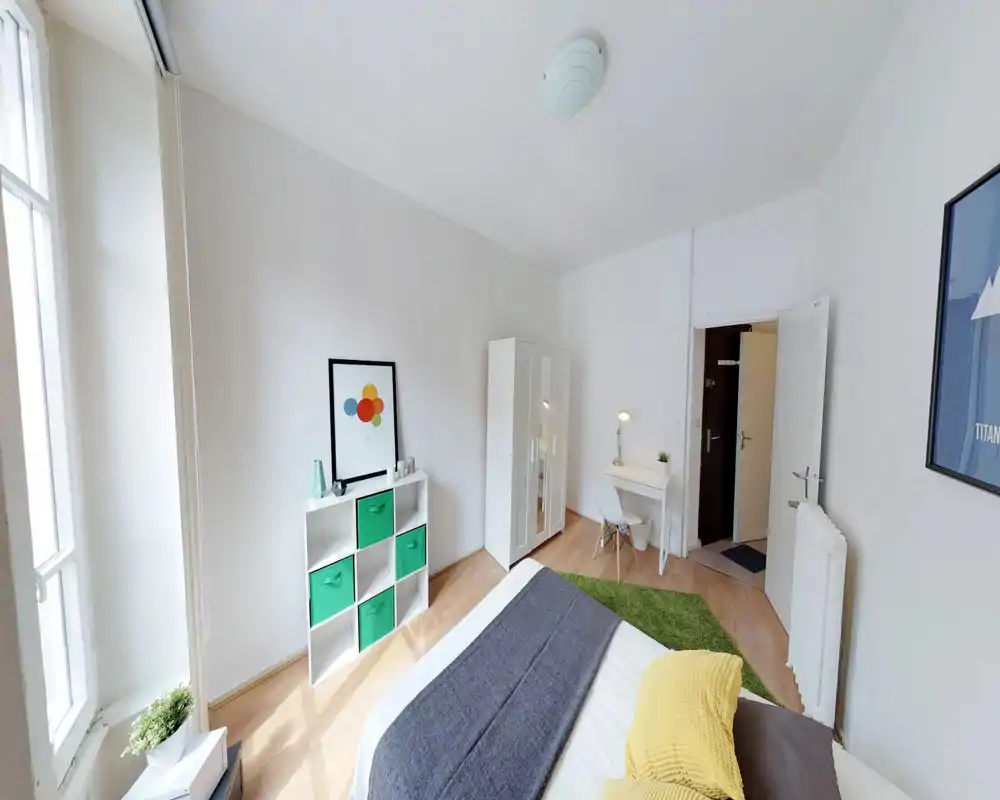 Bright, modern bedroom with a large bed dressed in neutral linens, colourful accent pillows and a green rug. The room features a wardrobe, desk with lamp, shelving with storage bins and a tall window that floods the space with daylight.
