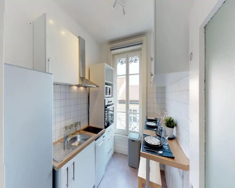 Compact, modern kitchen with white cabinets, wooden countertops and a large window that fills the space with natural light. Includes sink, stovetop, oven, dishwasher and a breakfast bar set for two—ideal for a functional city apartment.