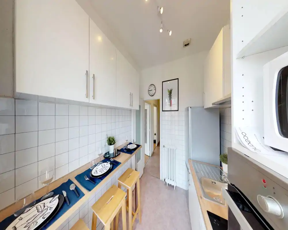 Bright modern galley kitchen with white cabinetry, tiled backsplash, compact sink and built-in appliances; a breakfast bar with wooden stools set for two offers a cozy dining spot.