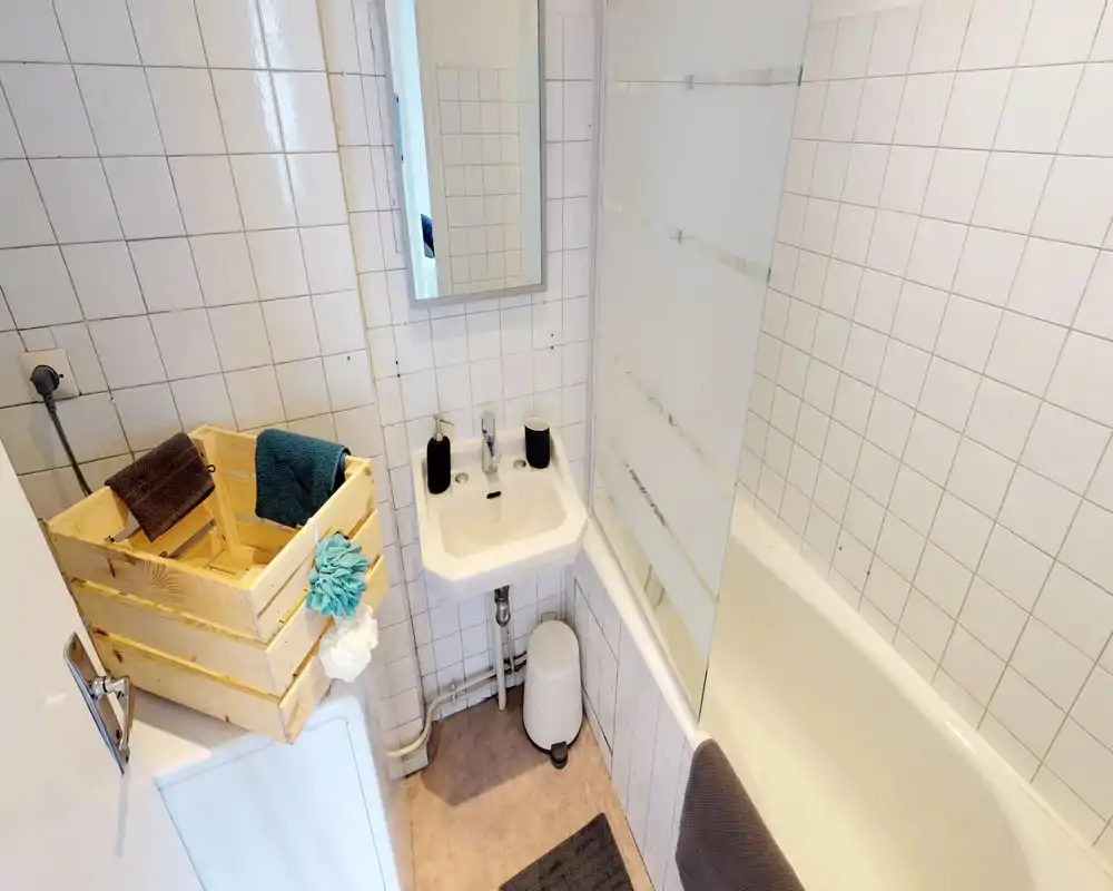 Compact, well-lit bathroom with a small sink, bathtub with glass screen, white tiled walls and simple accessories.