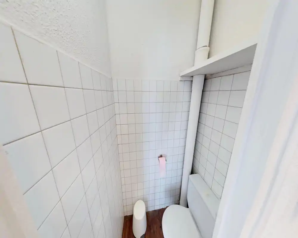 Compact toilet with white tiled walls, visible cistern and plumbing pipe; small waste bin and toilet paper holder included. Bright, utilitarian small bathroom suitable for a single occupant.