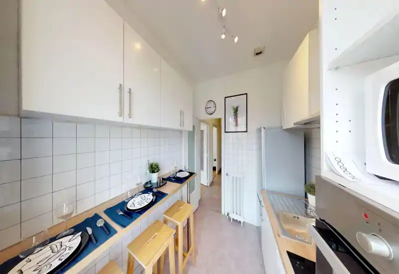 Bright modern galley kitchen with white cabinetry, tiled backsplash, compact sink and built-in appliances; a breakfast bar with wooden stools set for two offers a cozy dining spot.
