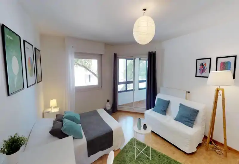 Bright, well-staged bedroom with a double bed, soft textiles and a small seating area by the window leading to a balcony.